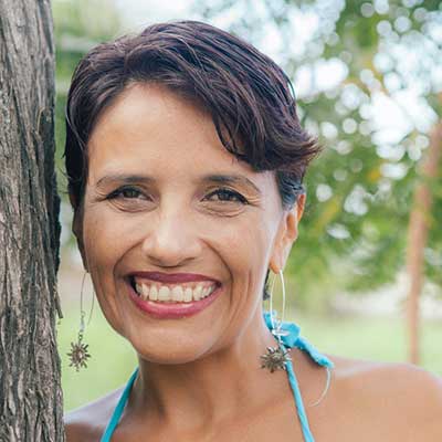 A smiling headshot of Paola Yepes leaning against a tree with a blurred, natural background.