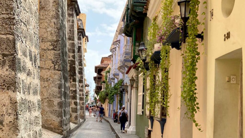 Narrow historic street in Cartagena, Colombia, with a massive ancient stone wall on the left and colorful colonial buildings with vine-covered balconies on the right.
