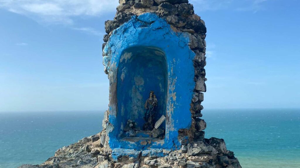 A rustic blue stone shrine with a religious statue overlooking the vast blue ocean under a clear sky at Cabo de la Vela, La Guajira, Colombia.