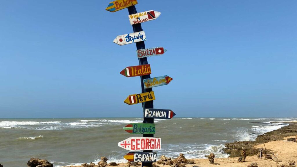 Multi-directional signpost with colorful arrows pointing to international cities and countries, standing on a sandy beach with the ocean in the background at Punta Gallinas, La Guajira, Colombia.