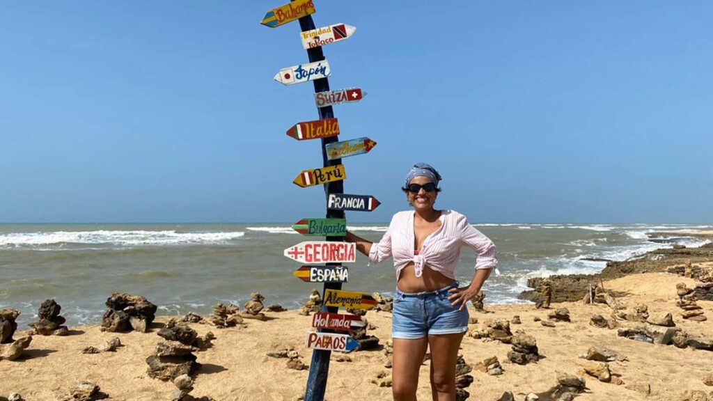Paola Yepes smiling and standing on a sandy beach at Punta Gallinas, Colombia, next to a signpost with arrows pointing to different international destinations.