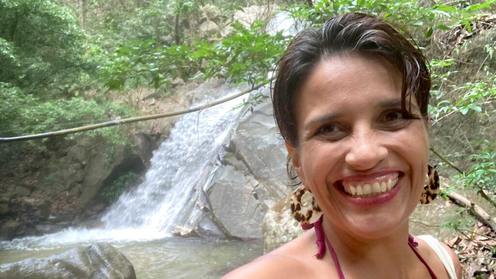 Paola Yepes smiling happily in front of a natural waterfall with lush green jungle in the background at Paso del Mango, Bonda, Colombia.