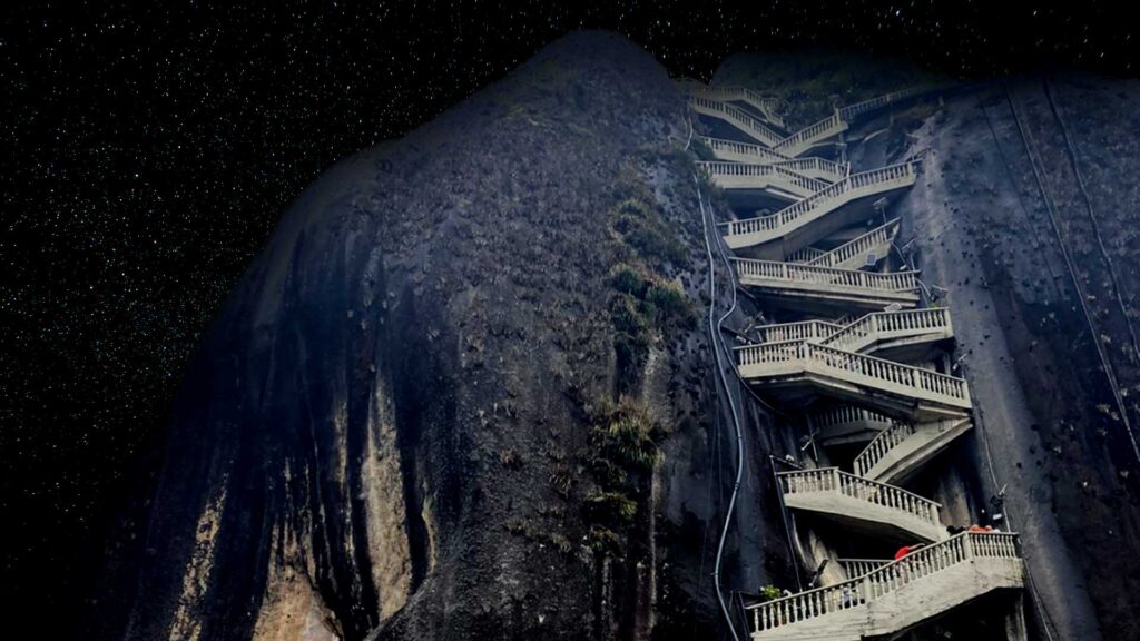 The iconic El Peñón de Guatapé rock in Colombia at night, with its zigzagging concrete staircase leading up the dark, massive stone face under a starry sky.
