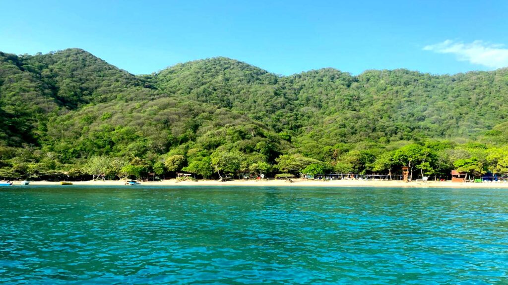 Wide view of Playa Cristal in Tayrona National Park, Colombia, showing vibrant turquoise water, a sandy beach, and dense green forested mountains under a clear blue sky.